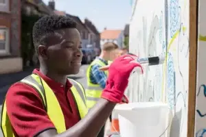 image of a man painting a mural outside in the foreground, other men can also be viewed helping in the background