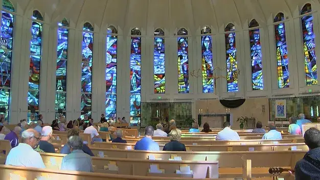 image of the inside of a church, the rows are filled with scattered people and stained glass windows are visible