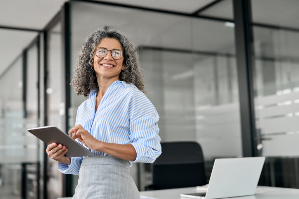 Smiling mature professional business woman holding tab looking away in office.