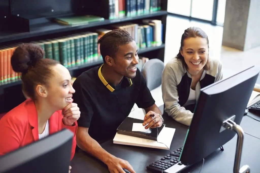 Image of three people looking at the center mans computer, behind them are bookshelves