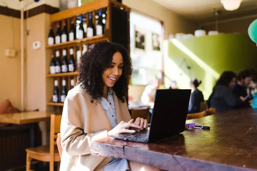 Image of a woman within a winery sitting at a table, smiling while working on her laptop alone