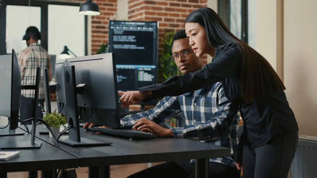 Two programmers holding laptop with coding interface walking towards desk and sitting down