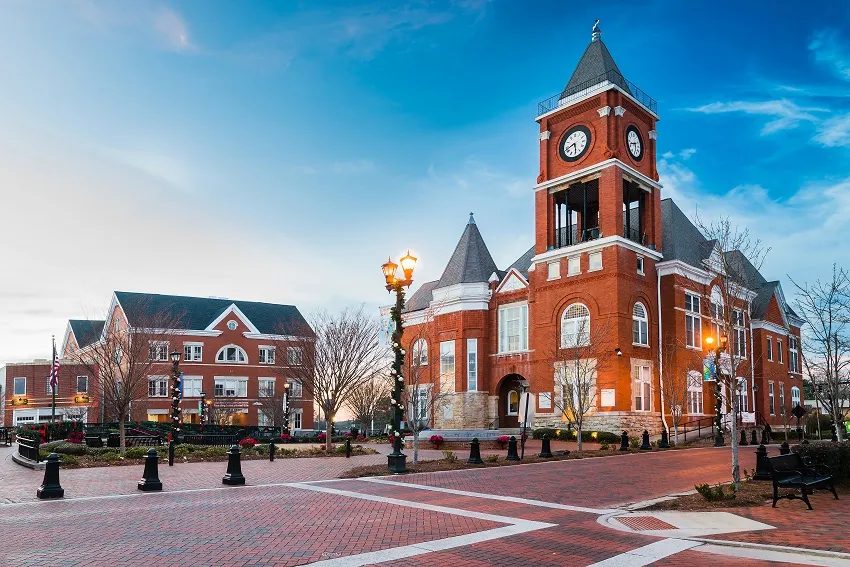Historic red brick clock tower building with a decorative clock, flanked by modern structures, set against a vibrant blue sky