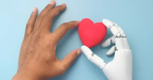 A human hand and a robotic hand delicately hold a red heart against a light blue background, symbolizing connection and empathy