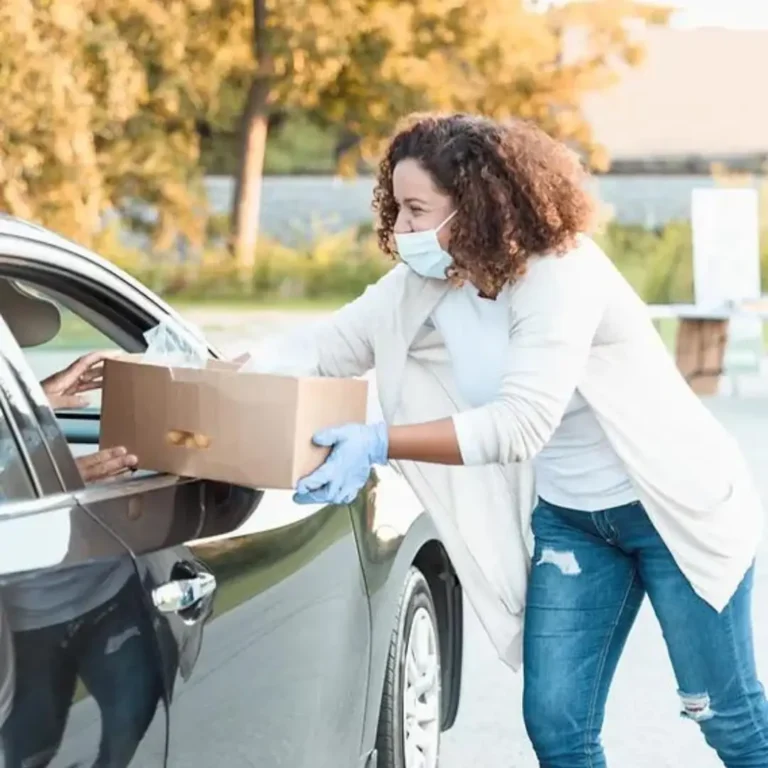 Image of a woman in jeans with a mask and gloves handing a box of items through a passenger door window of a car